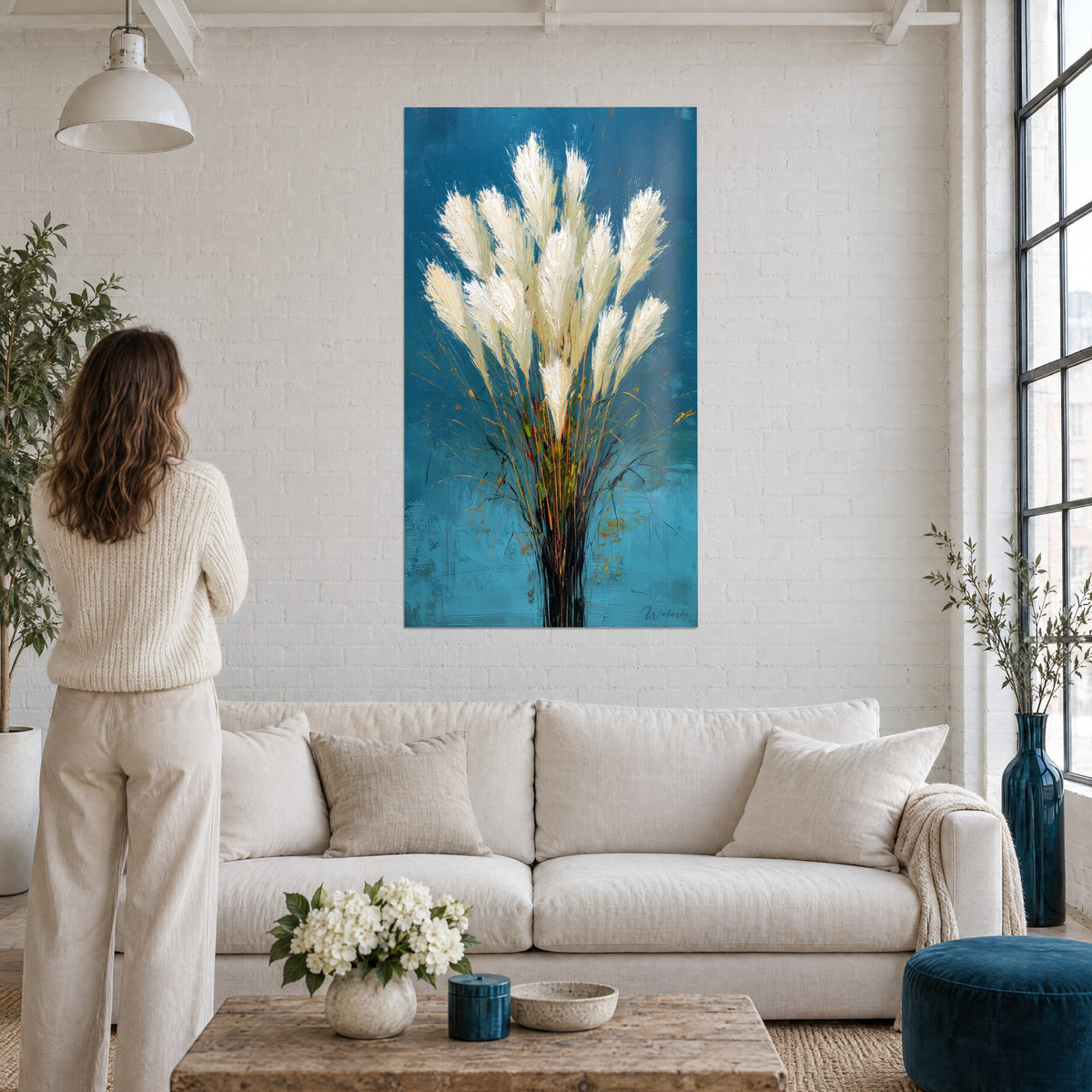 Schilderij crème pampasgras met dikke pluimen op marineblauwe achtergrond, vrouw van achteren, boven een bank in een lichte woonkamer