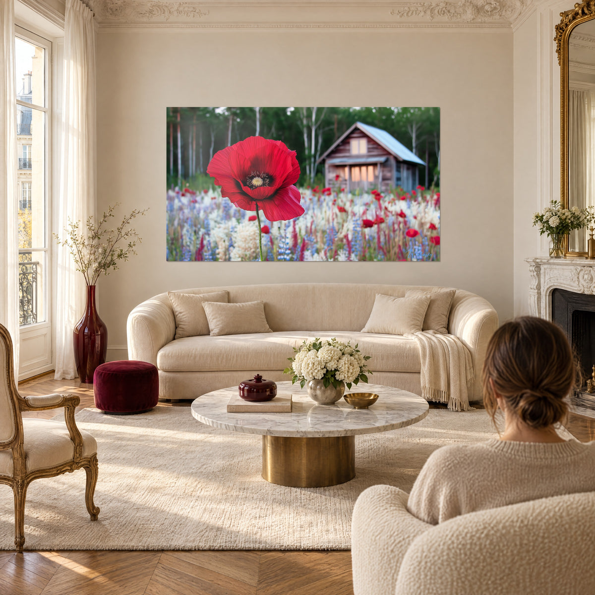 Wall art of a giant carmine red poppy in front of a wooden chalet, blue and white flower meadow, woman seen from behind above a sofa in a bright living room