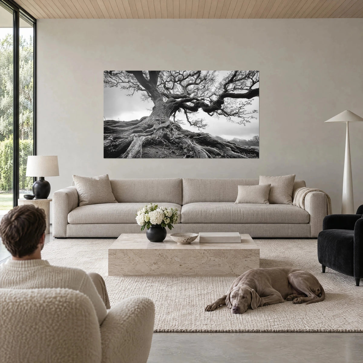 Quadro di una quercia secolare dai rami contorti, cielo grigio perla e radici color carbone, una donna e il suo Weimaraner sopra un divano in un soggiorno luminoso