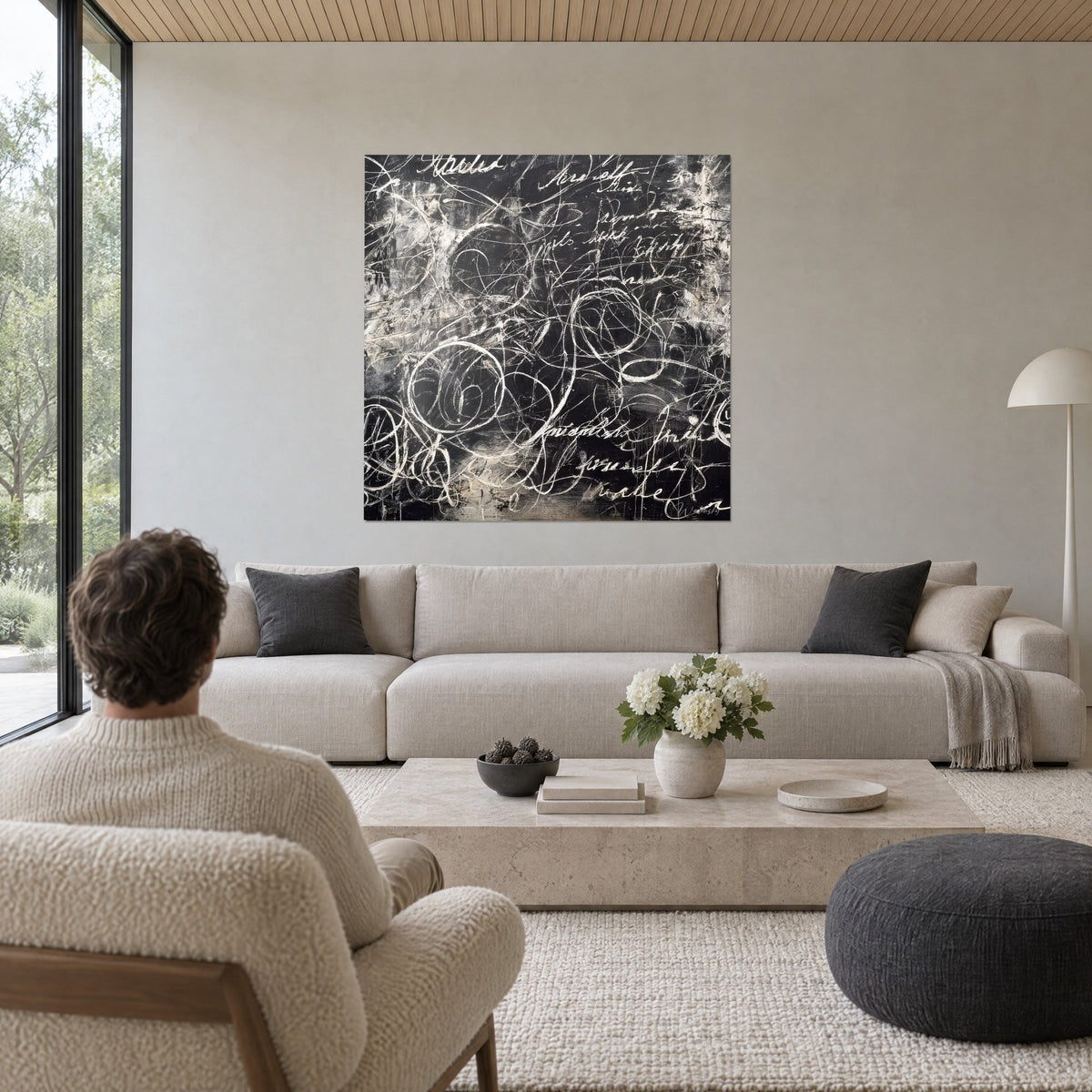 Wall art with gestural calligraphy in cream white on a black background, loops and chalk lines, man seen from behind, above a sofa in a bright living room