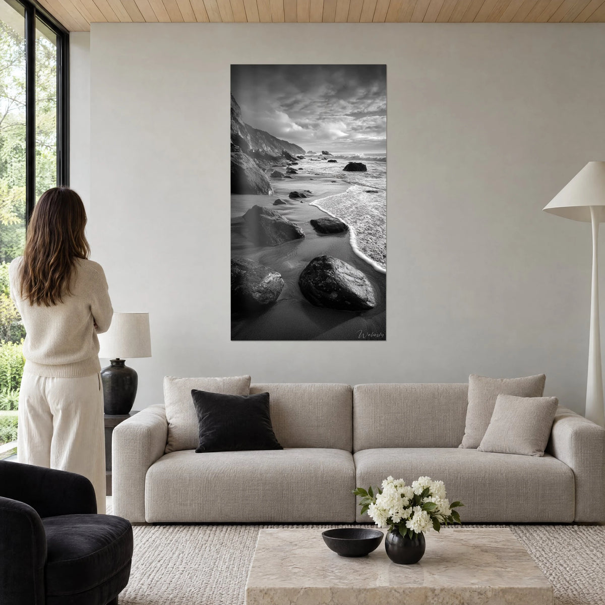 Tableau plage rocheuse en noir et blanc, galets sombres et écume blanche, femme de dos, au-dessus d'un canapé dans un salon lumineux