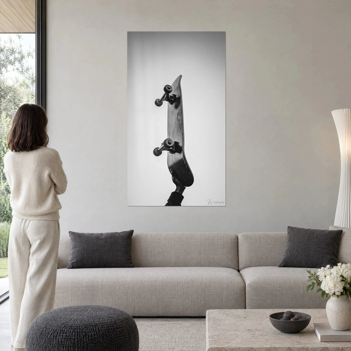 Quadro skateboard sollevata verso il cielo, ruote nere e grip grigio, donna di spalle, sopra un divano in un soggiorno luminoso