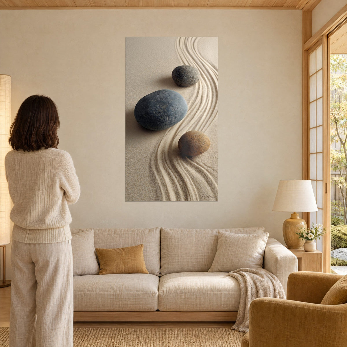 Wall art with three blue-grey and ochre pebbles on beige sand striped with waves, a woman seen from behind, above a sofa in a bright living room
