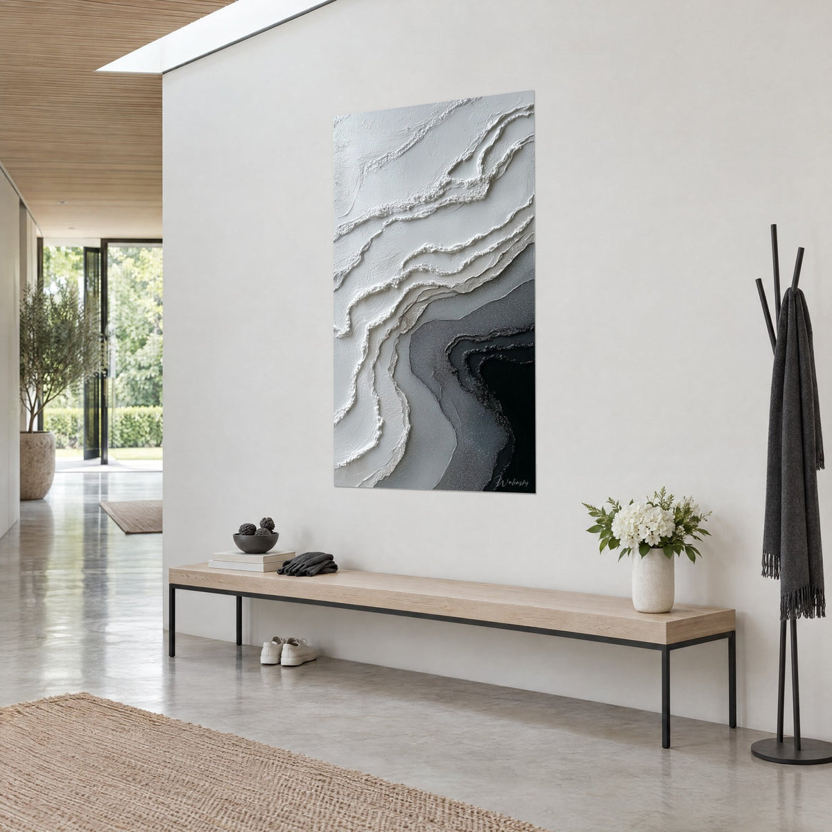 Quadro con strati minerali in rilievo color crema e antracite, appeso a una parete chiara accanto a una panca in legno e a un mazzo bianco, ingresso luminoso
