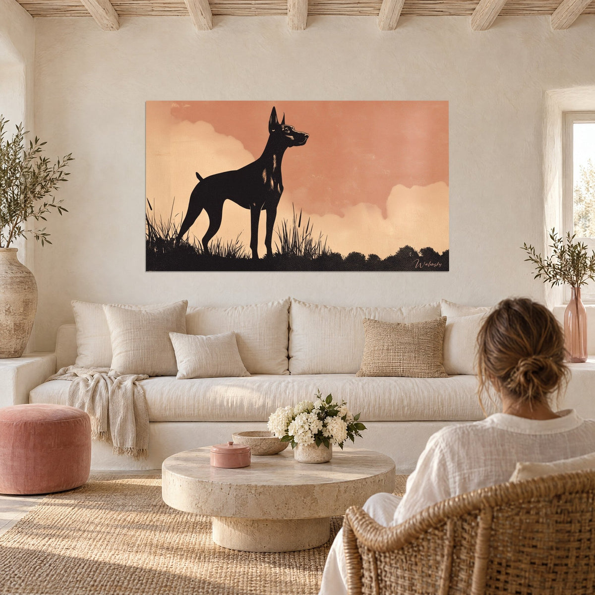 Gemälde Dobermann als schwarze Silhouette vor rosa-lachsfarbenem Himmel und cremeweißen Wolken, über einem Sofa in einem hellen Wohnzimmer