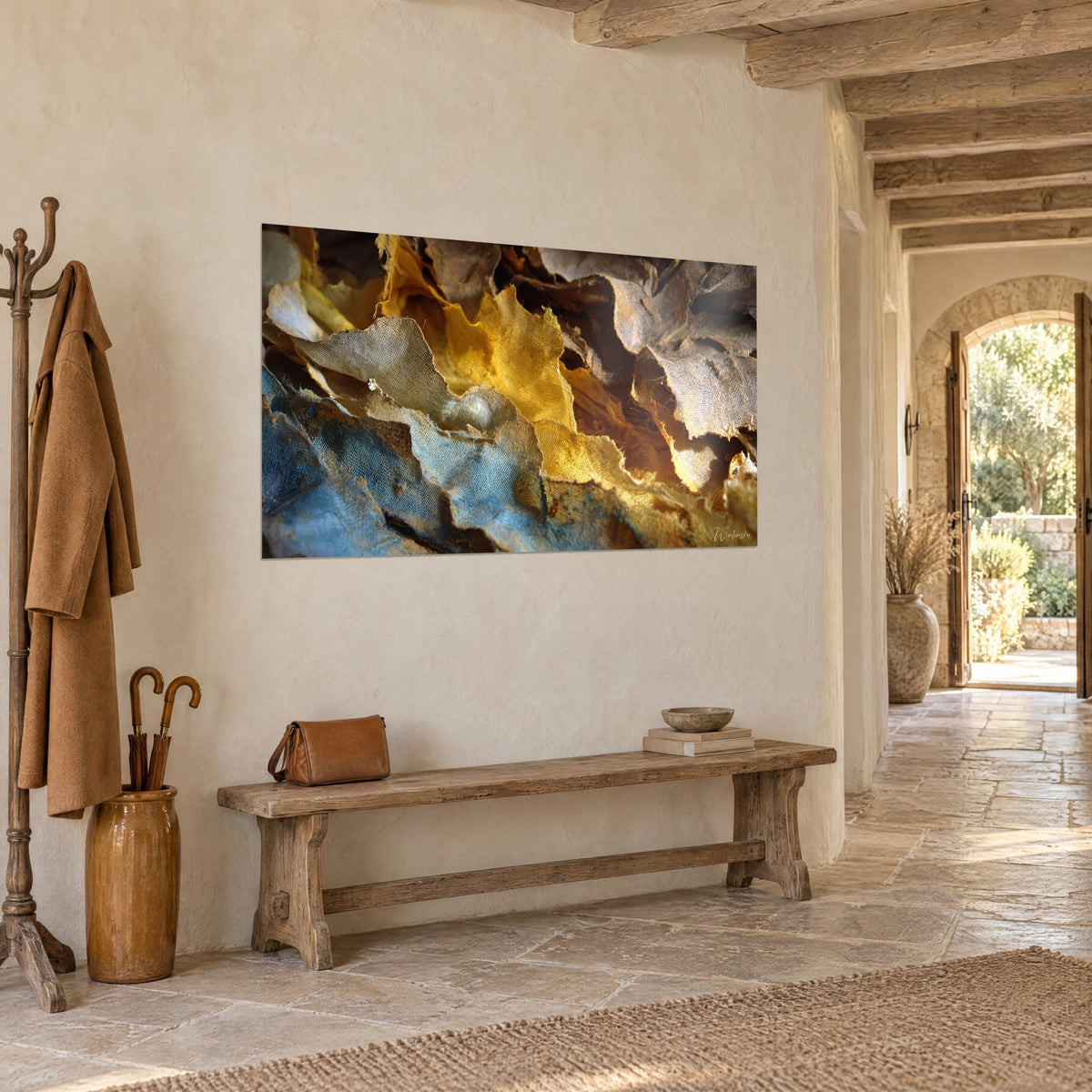 Wall art with ochre yellow and tawny brown linen rags with fringed edges, seen three-quarter view in the entrance of a stone house