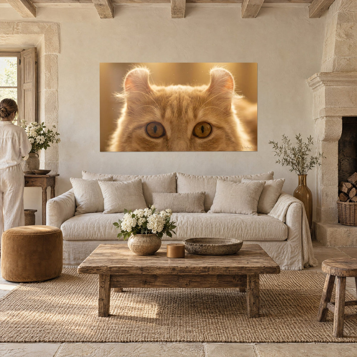 Wall art of a ginger cat with curled ears, amber eyes and honey-toned background, hung across from two visitors above a sofa in a bright living room