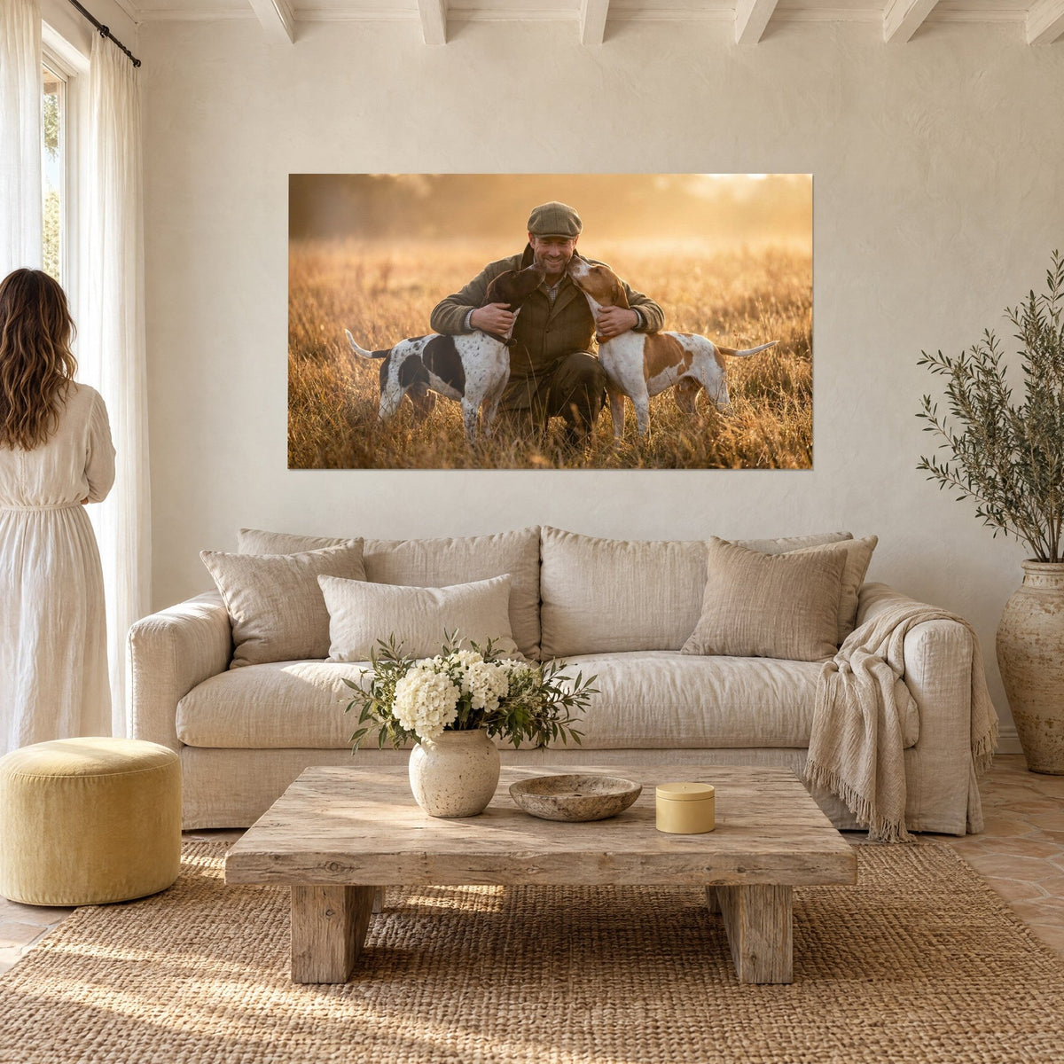 Quadro cacciatore con berretto cachi accucciato tra due cani, erbe ambrate e dorate, sopra un divano in un soggiorno luminoso