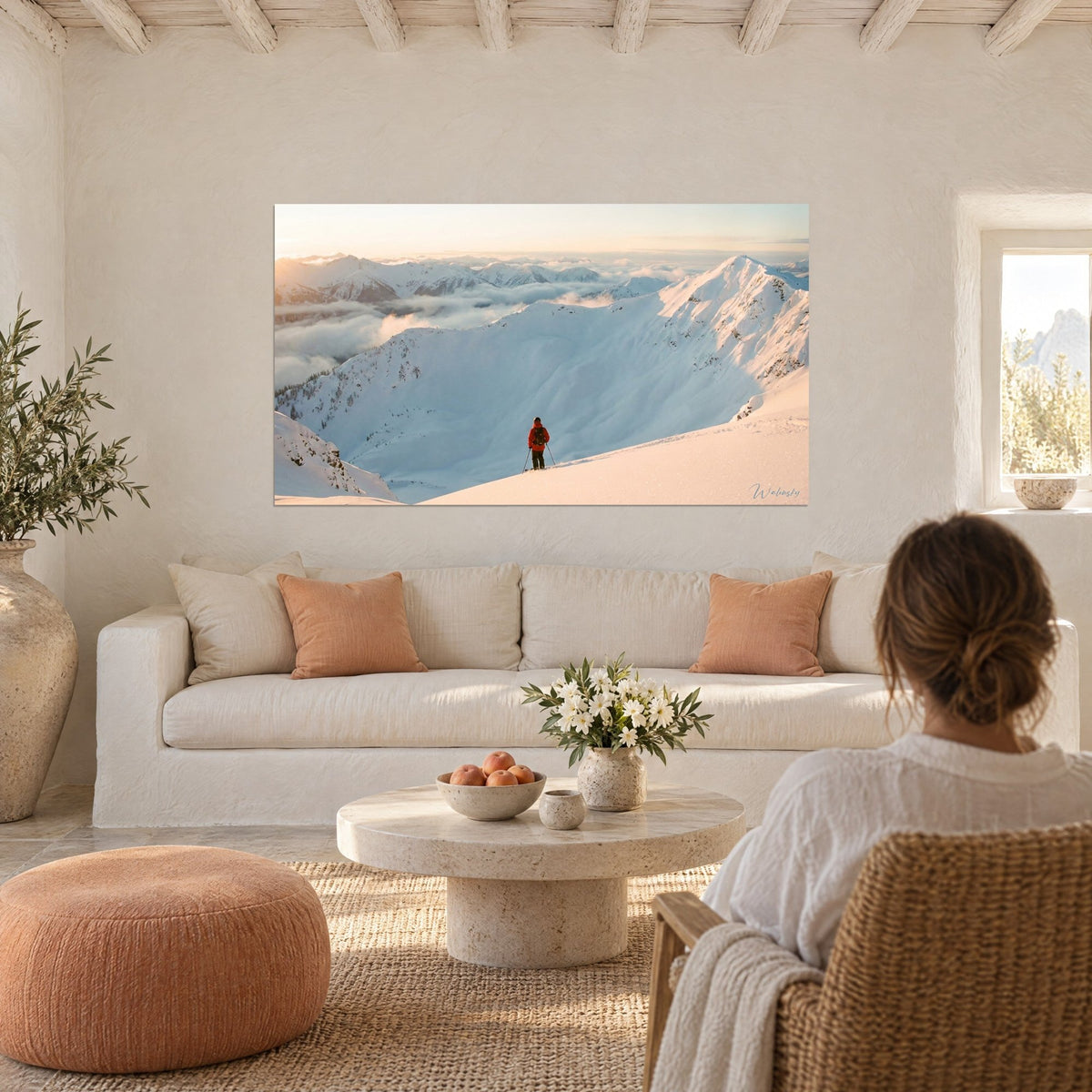 Gemälde Skifahrer in orangefarbener Jacke vor einem verschneiten Zirkus und einem rosa Wolkenmeer, über einem Sofa in einem hellen Wohnzimmer