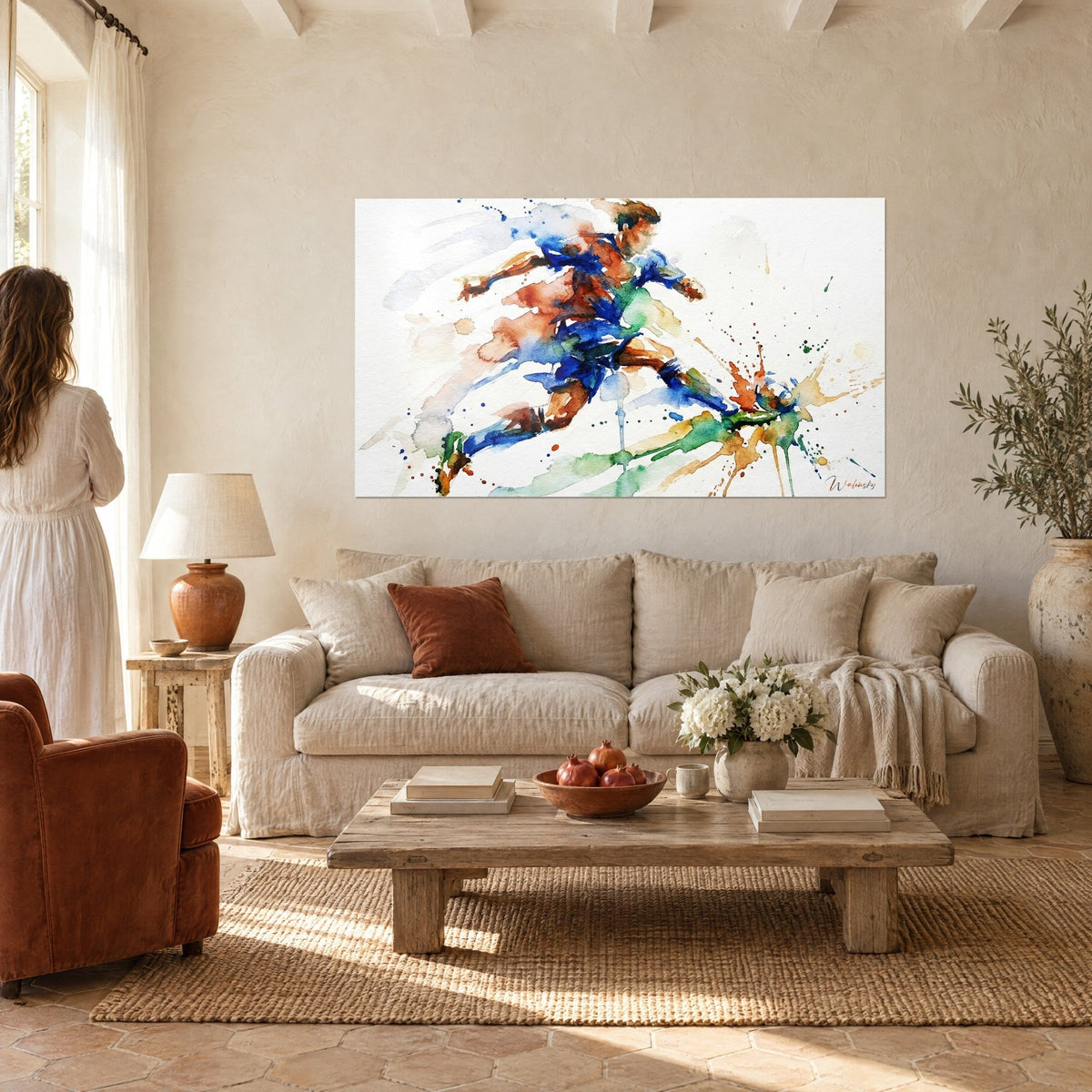 Schilderij van een voetballer in volle sprint, kobaltblauw shirt met roestrode vlekken, tegen een crèmekleurige muur, vrouw van achteren, boven een bank in een lichte woonkamer