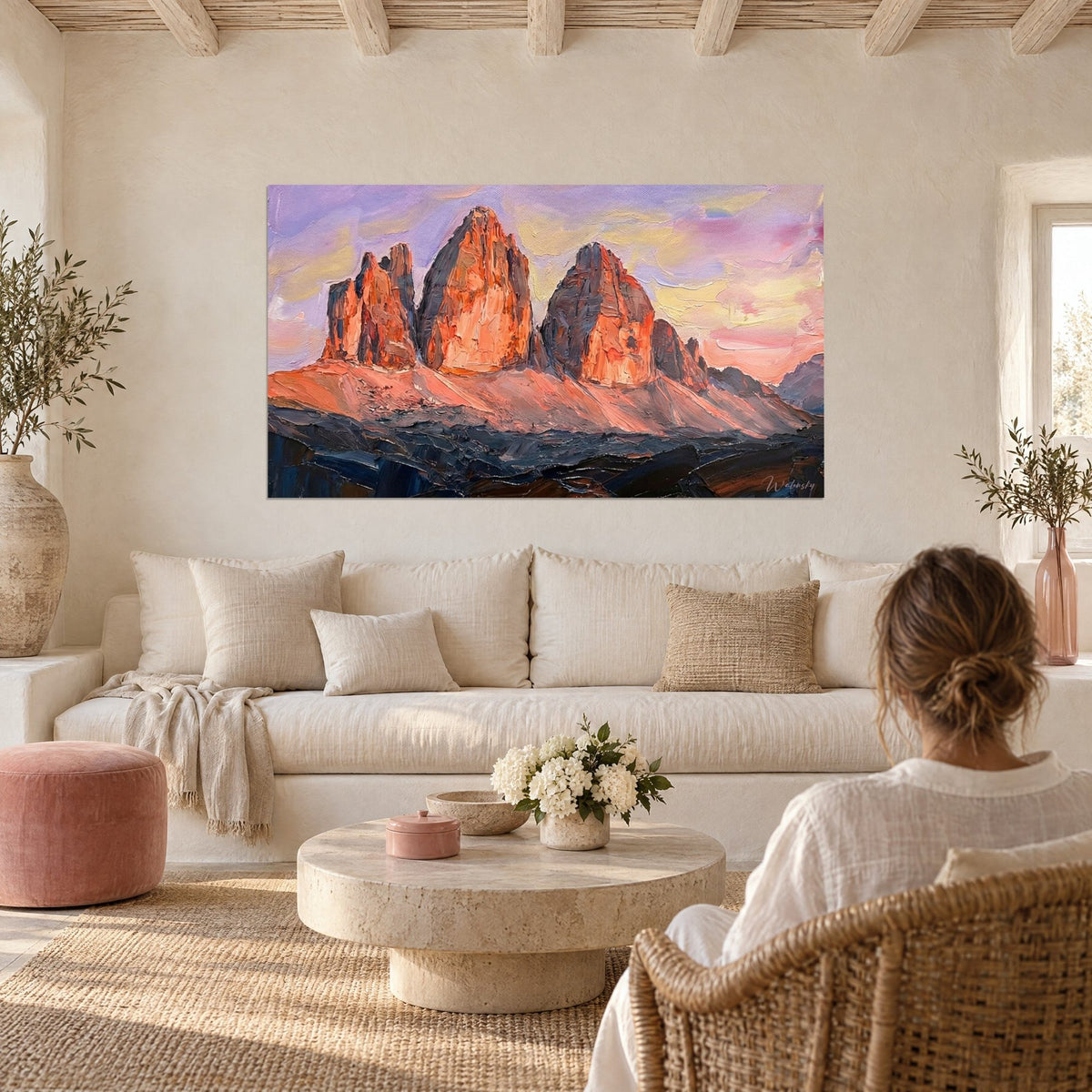 Tableau Tre Cime de Lavaredo en flammes orange sur ciel lilas, une femme de dos le regarde au-dessus d'un canapé dans un salon lumineux