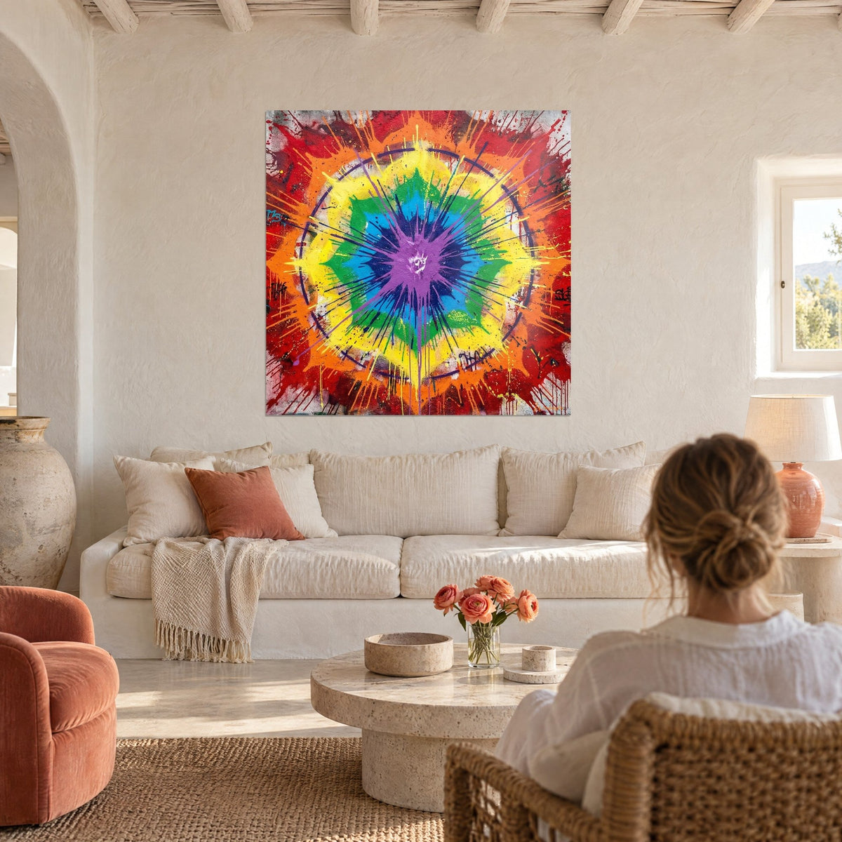 Wall art with an explosive mandala of yellow and green petals and a splattered violet centre, facing a woman seen from behind, above a sofa in a bright living room