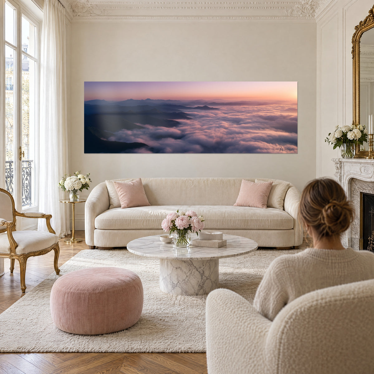 Schilderij Pyreneese wolkenzee in roze en mauve onder een oranje hemel, koppel van achteren, boven een bank in een lichte woonkamer