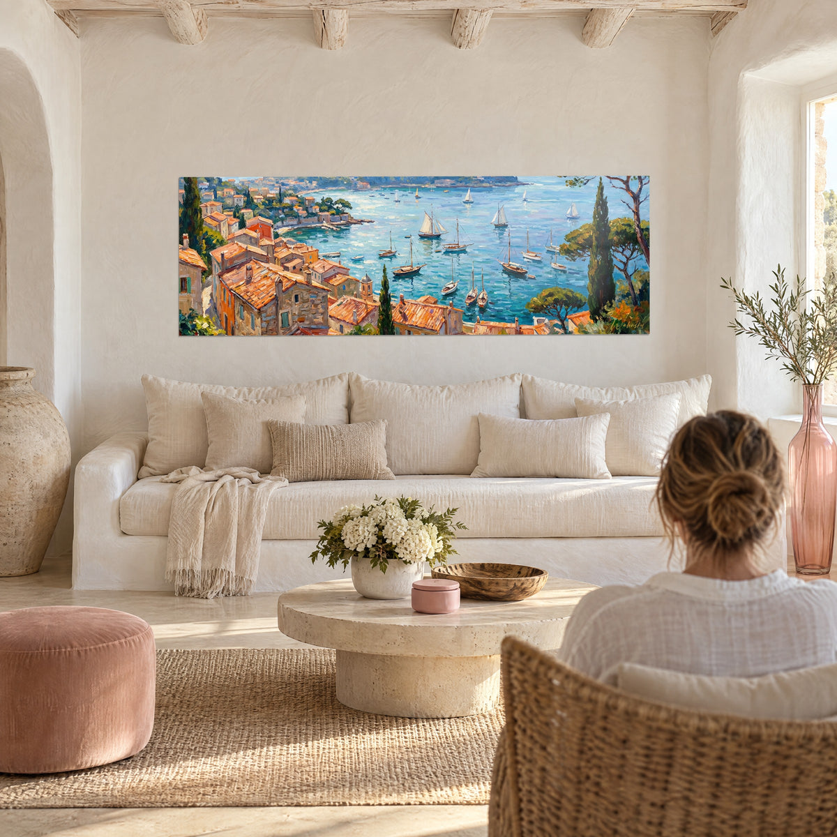 Gemälde Mittelmeerbucht mit weißen Segelbooten, orangefarbenen Dächern und Zypressen, eine Frau von hinten über einem Sofa in einem hellen Wohnzimmer
