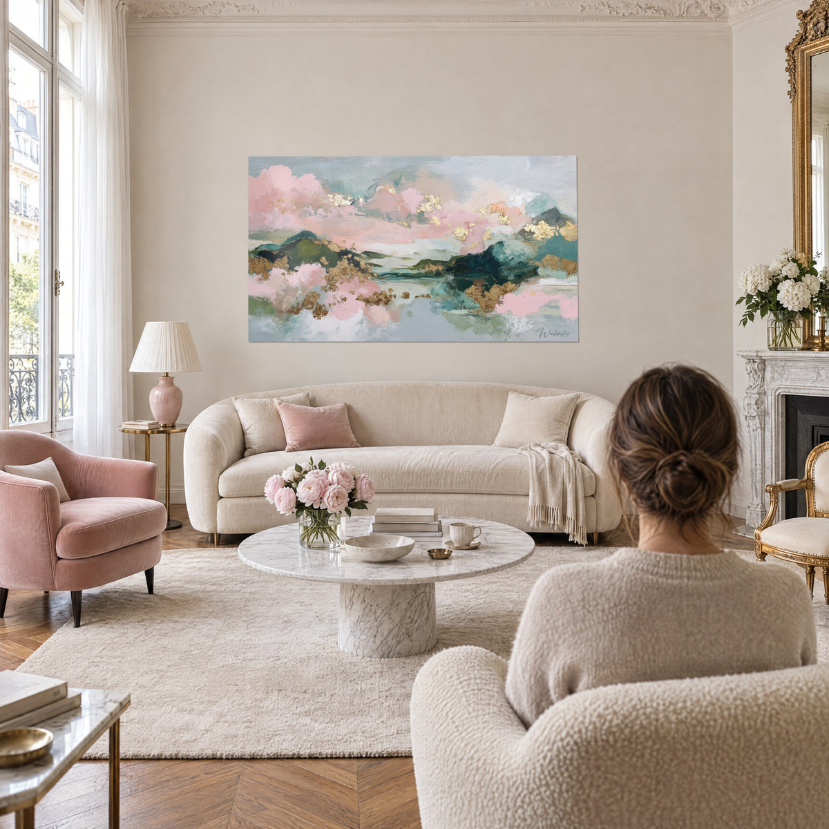 Schilderij pastelkleurige bergen in een poederroze gloed met turquoise reflecties en gouden glinsteringen, vrouw van achteren boven een bank in een lichte woonkamer
