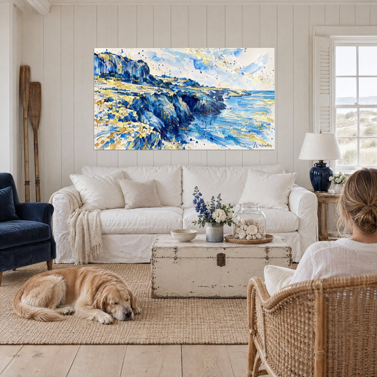 Tableau falaise rocheuse en aquarelle bleu cobalt et or, bateau blanc sur mer calme, retriever couché, au-dessus d'un canapé dans un salon lumineux