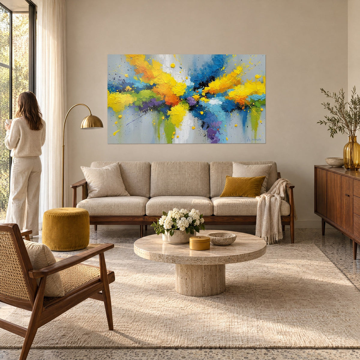 Wall art with splashes of yellow and blue paint on a grey background, woman seen from behind above a sofa in a bright living room