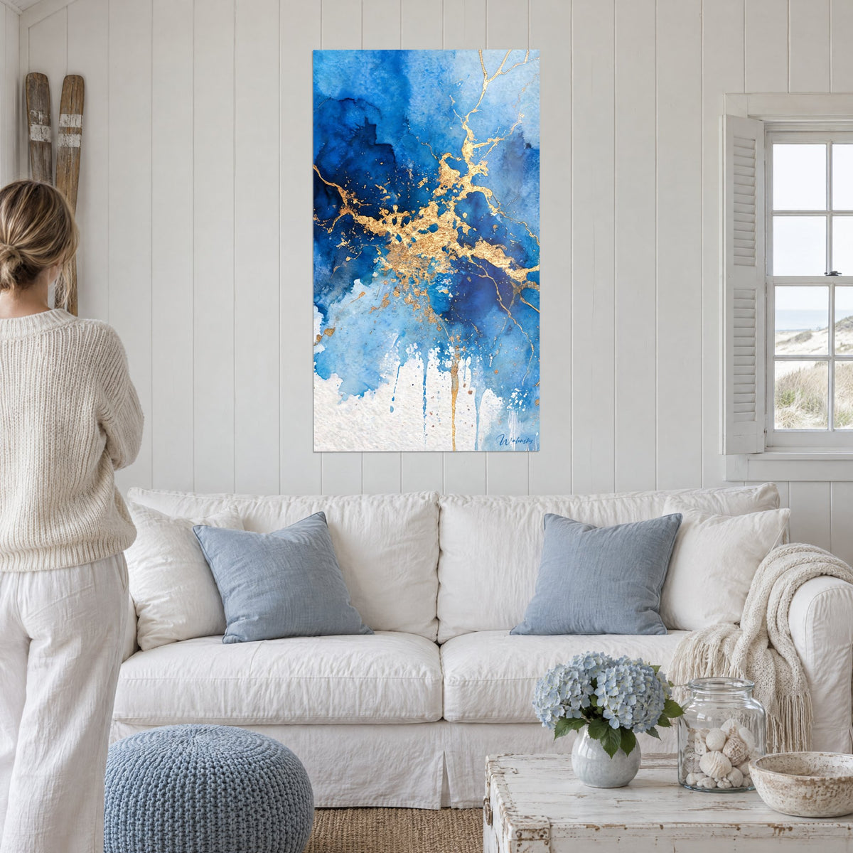 Tableau aquarelle indigo fissurée de veines dorées, nuages cyan et bas blanc, au-dessus d'un canapé dans un salon lumineux