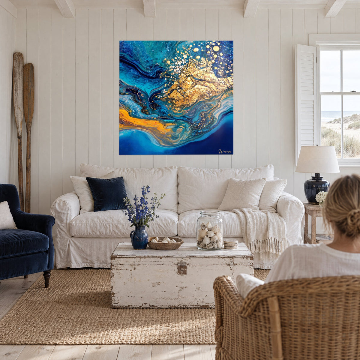Tableau marbrures turquoise et bleu nuit striées de paillettes dorées, une femme de dos, au-dessus d'un canapé dans un salon lumineux