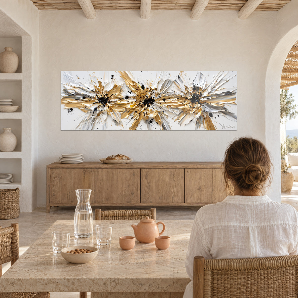 Schilderij metalen uitbarsting van goud en gebroken wit, dikke impasto-streken, tegenover een vrouw die aan tafel zit in een gekalkte woonkamer