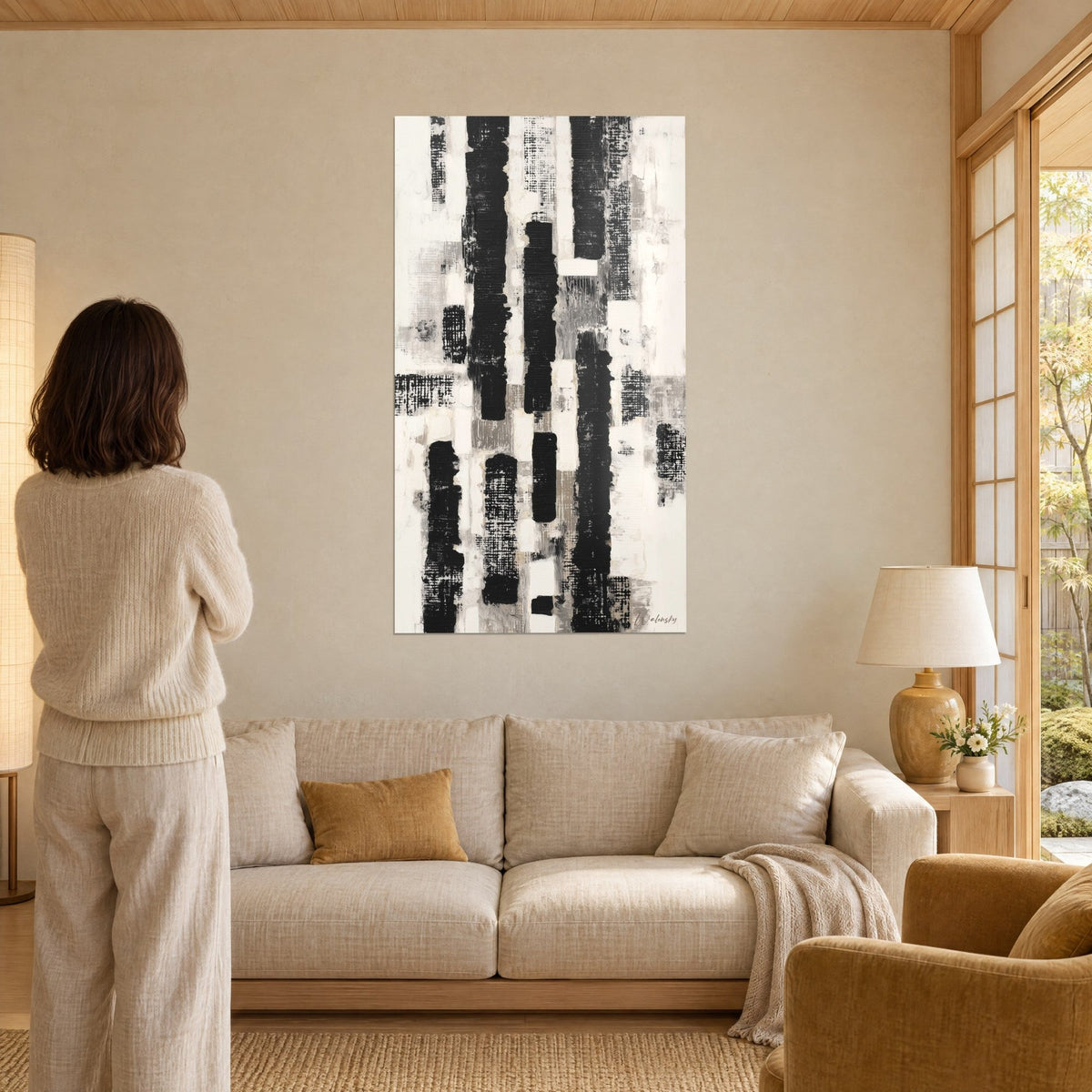 Quadro astratto in bianco e nero con colonne verticali testurizzate, toni grigi e crema, donna di spalle sopra un divano in un soggiorno luminoso