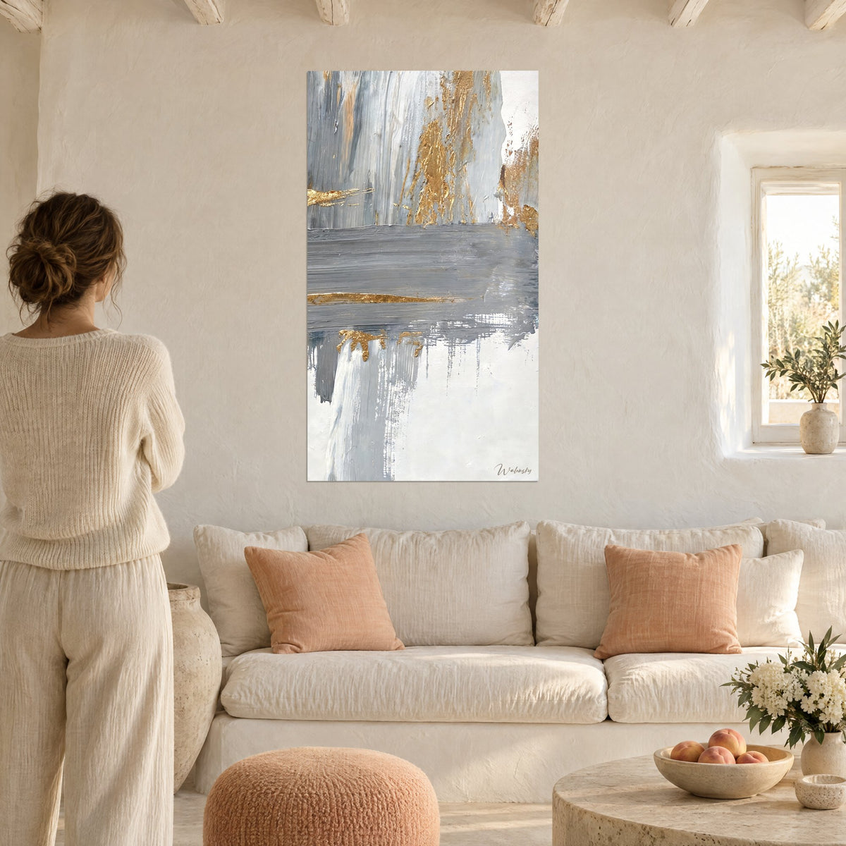 Tableau abstrait gris perle traversé de feuilles d'or, femme de dos en pull crème, au-dessus d'un canapé dans un salon lumineux