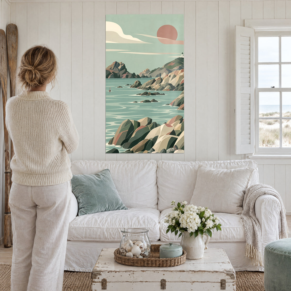 Quadro costa rocciosa sotto un sole terracotta, mare verde acqua, faro bianco, donna di spalle sopra un divano in un soggiorno luminoso