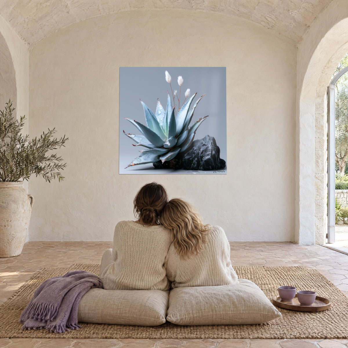 Tableau aloès bleu pervenche gorgé de rosée et roche anthracite, mur blanc voûté, deux personnes enlacées de dos sur des coussins