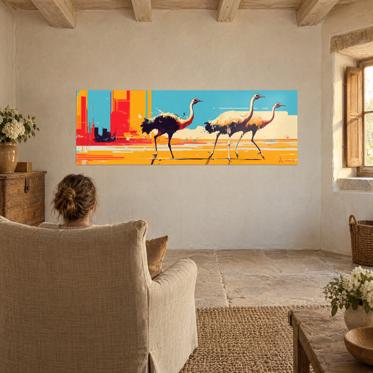 Wall art trio of large striding birds, rosy necks, bright yellow ground and blue sky, facing a linen armchair in a stone living room