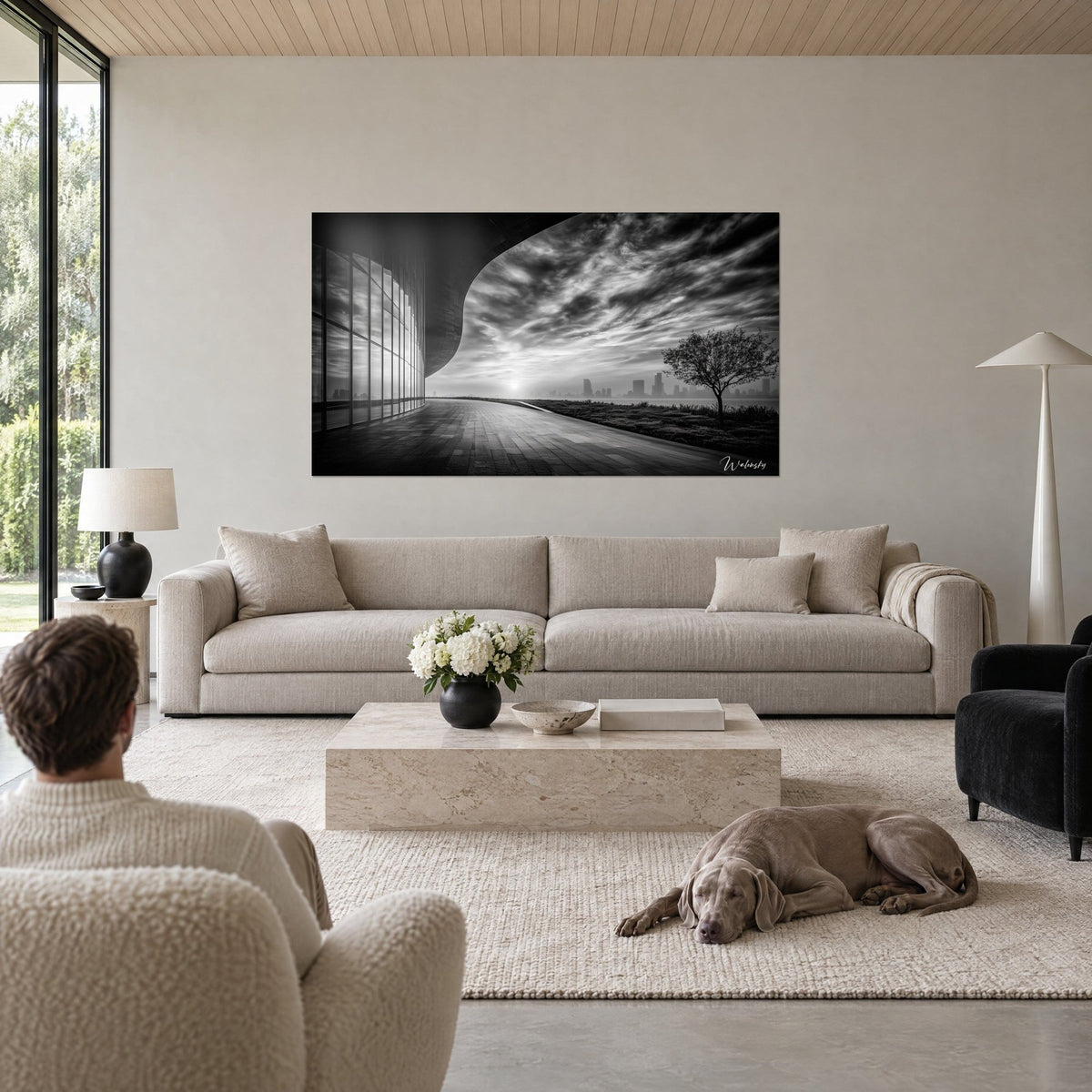 Tableau façade vitrée incurvée sous un ciel nuageux noir et blanc, arbre solitaire, homme de dos au-dessus d'un canapé dans un salon lumineux