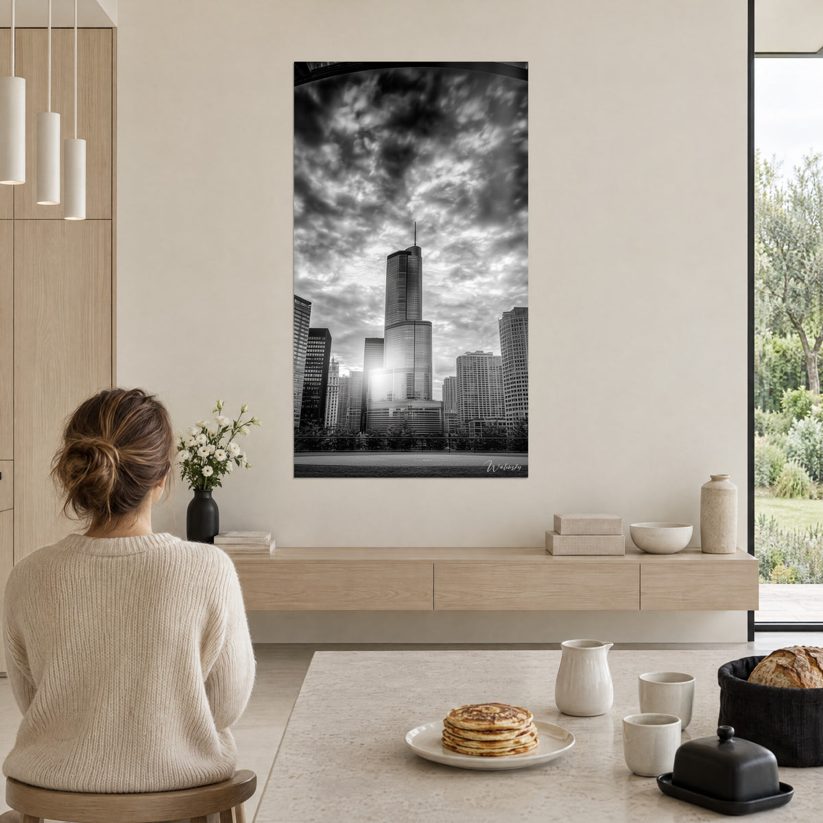Wall art of a monochrome skyline under a stormy sky, deep blacks and vivid whites, woman seen from behind with a loose bun, hung above a breakfast table