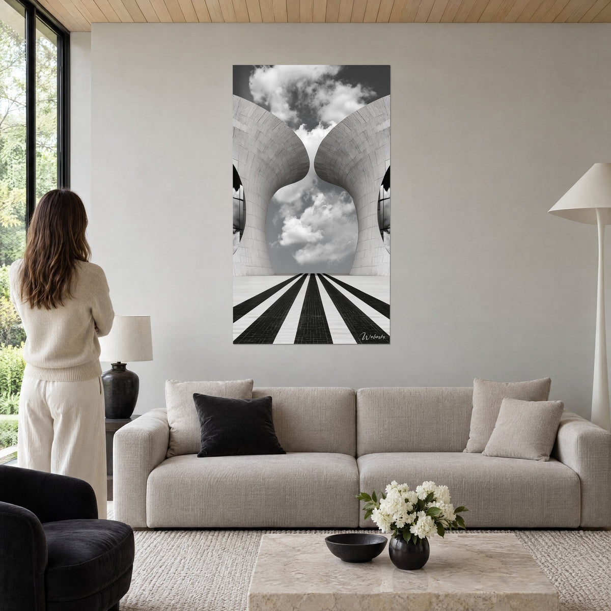Schilderij tweelingtorens van witte steen in elkaar verstrengeld onder een bewolkte hemel, zwart-wit gestreepte vloer, vrouw van achteren, boven een bank in een lichte woonkamer