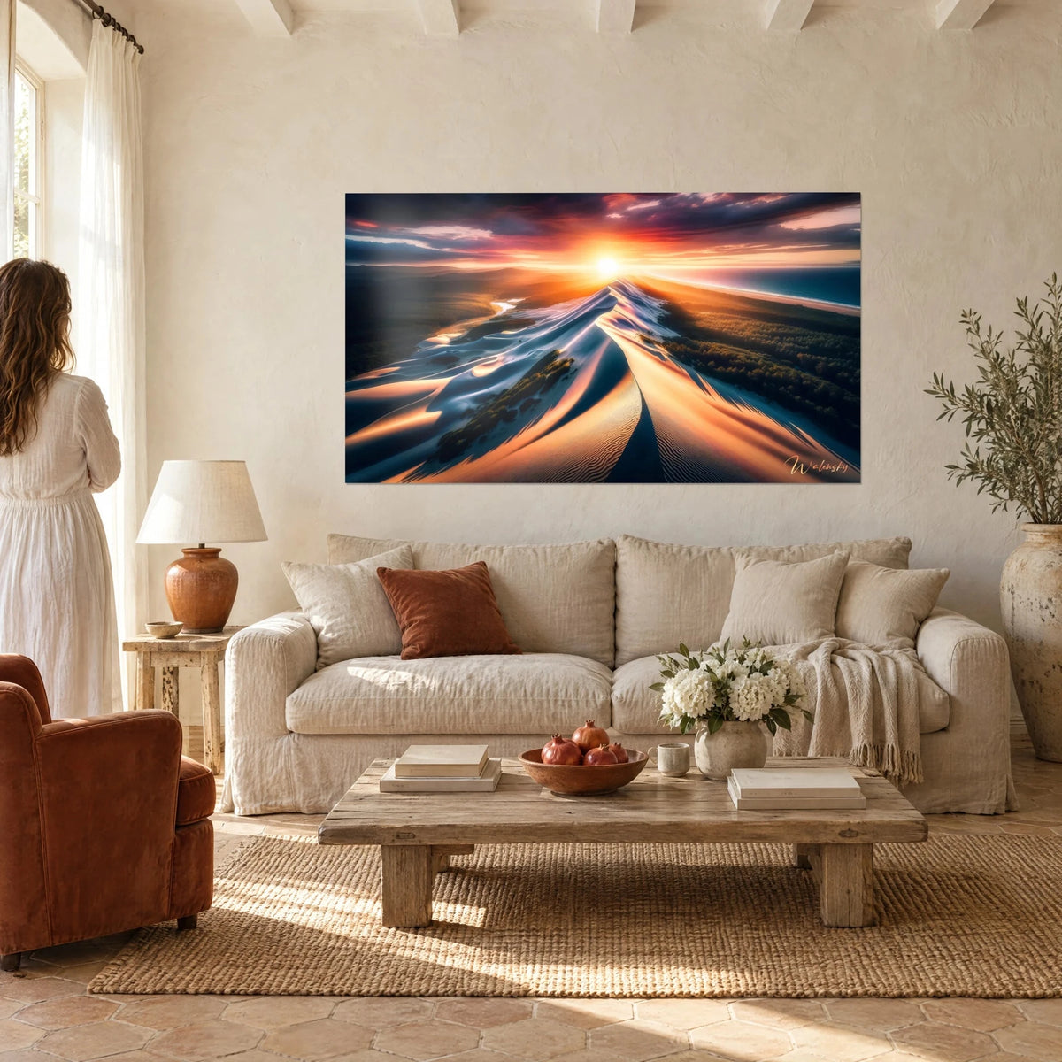 Quadro di una cresta di duna scolpita da un sole arancio e rosa, ombre azzurrate, vista di una donna di spalle sopra un divano in un soggiorno luminoso