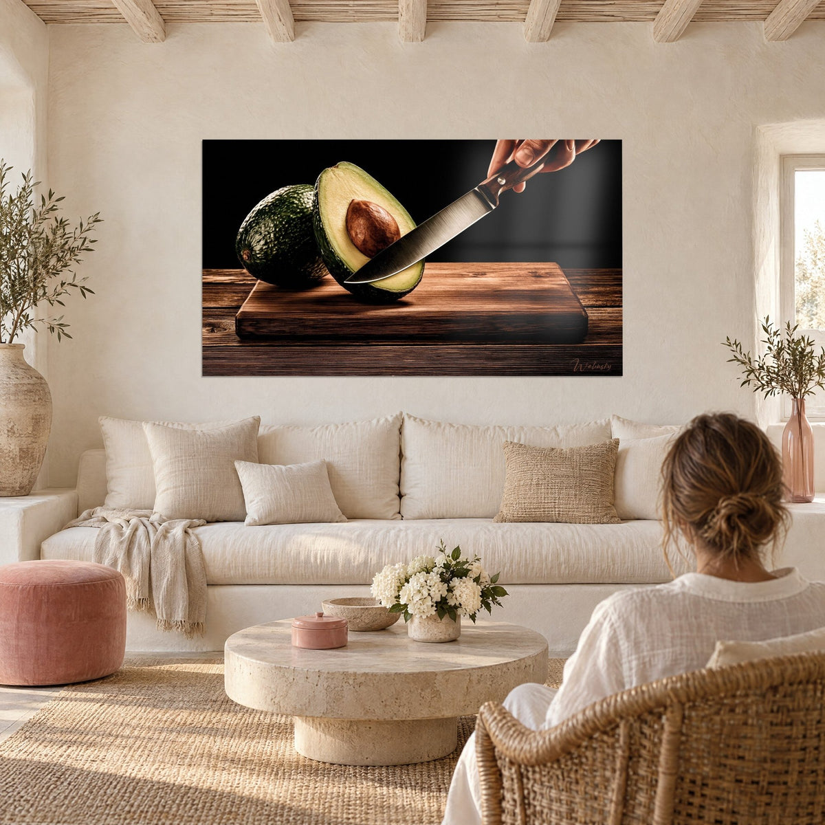 Wall art half avocado with brown pit, steel knife resting on amber oak board, hung above a sofa in a bright living room