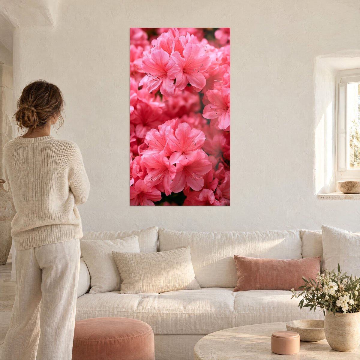 Quadro azalee rosa corallo in primo piano, petali spiegazzati e stami sottili, donna di spalle, sopra un divano in un soggiorno luminoso