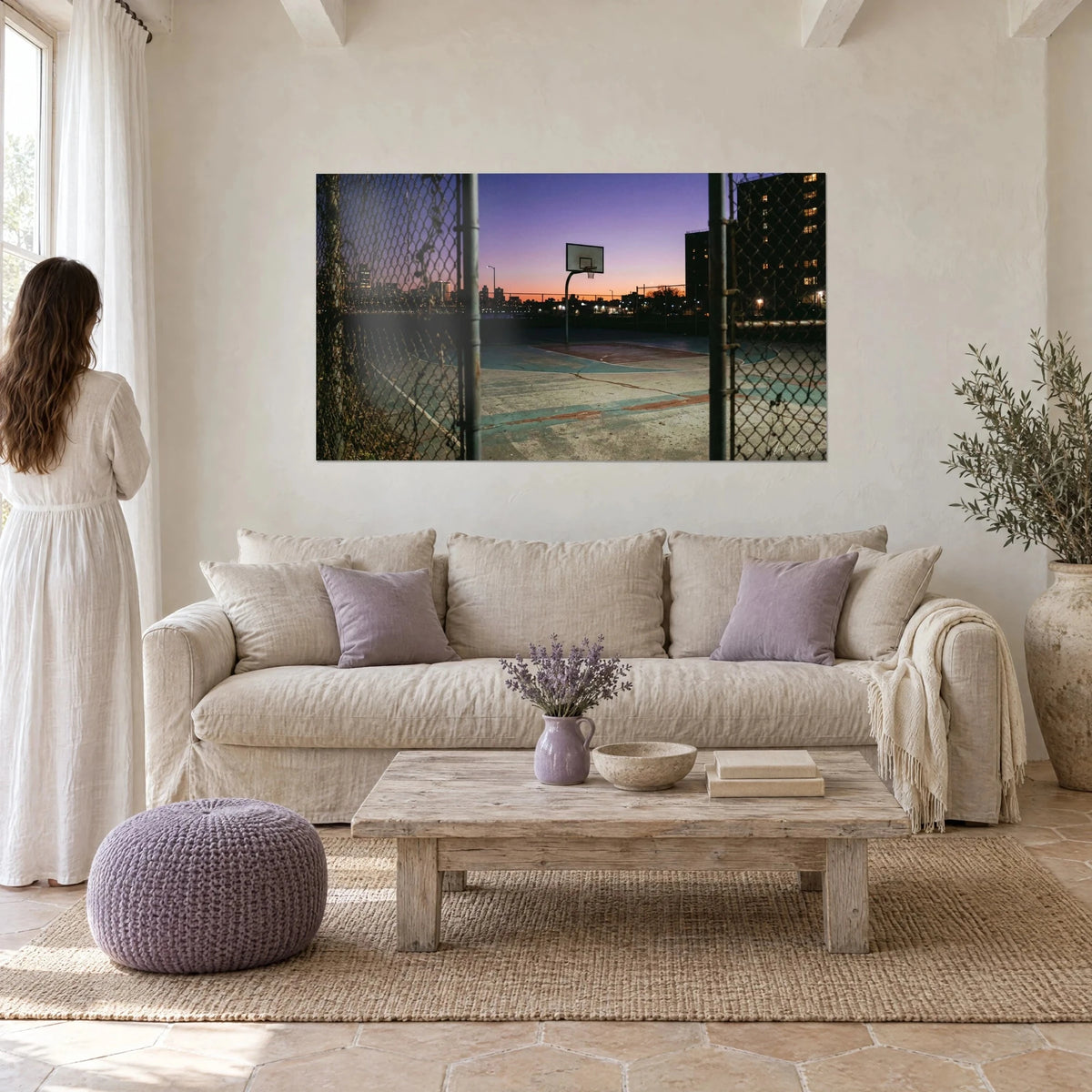 Gemälde Basketballfeld mit Maschendrahtzaun unter violettem und orangefarbenem Himmel, Frau von hinten, über einem Sofa in einem hellen Wohnzimmer