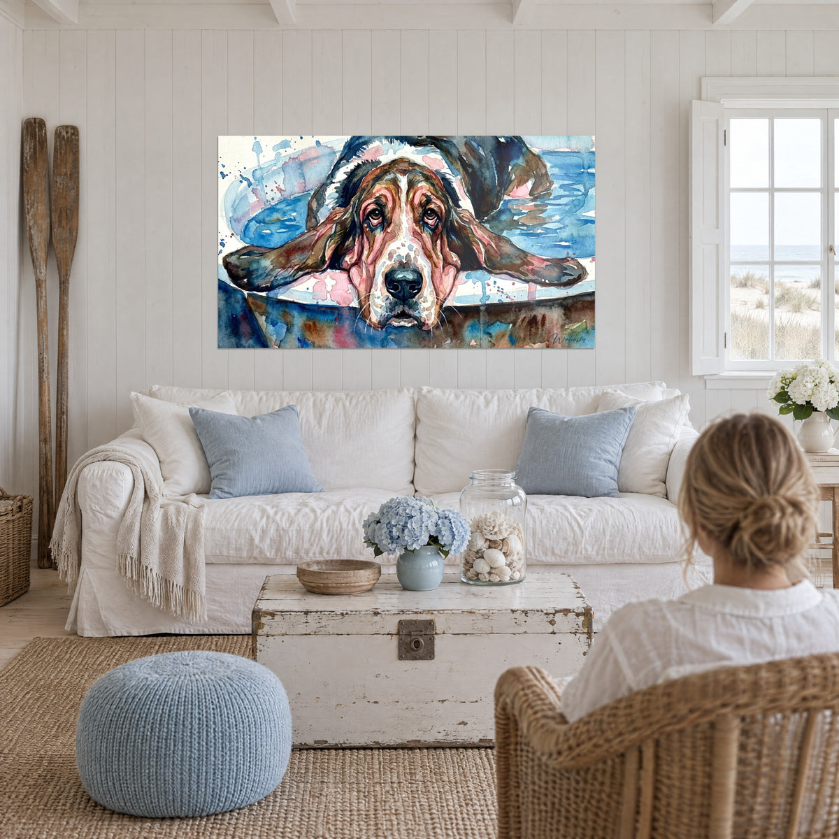 Cuadro de un basset hound con orejas largas y marrones y mirada melancólica, fondo azul acuarela, sobre un sofá en un salón luminoso