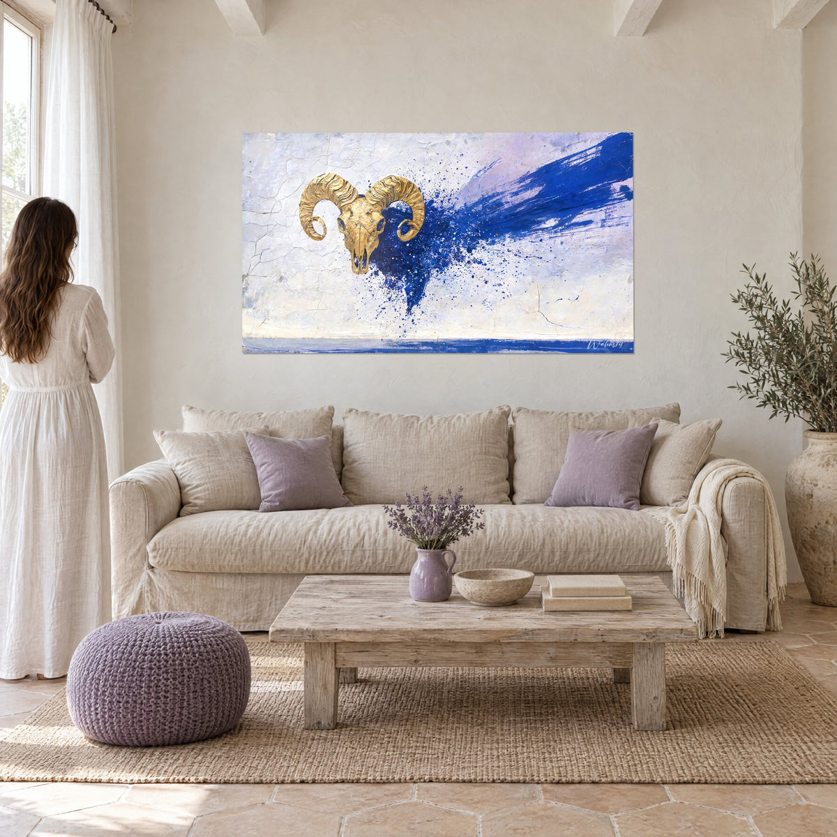 Tableau crâne de bélier doré aux cornes torsadées, éclaboussures bleu marine sur fond blanc craquelé, au-dessus d'un canapé dans un salon lumineux