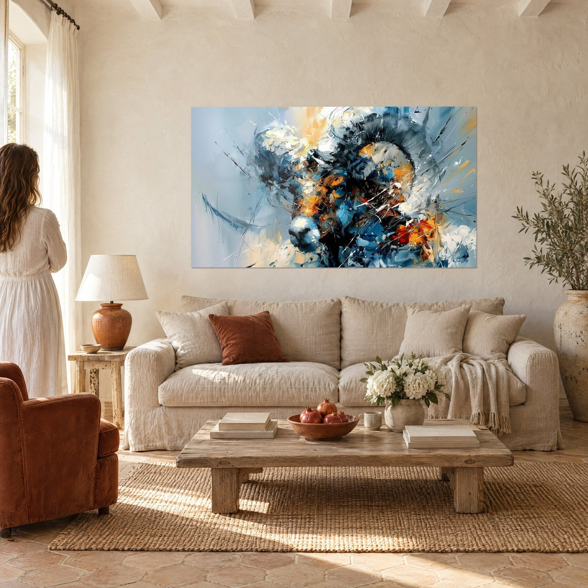 Quadro ariete dalle corna ritorte grigio ardesia, schegge ocra e tocchi rossi, donna di spalle, sopra un divano in un soggiorno luminoso