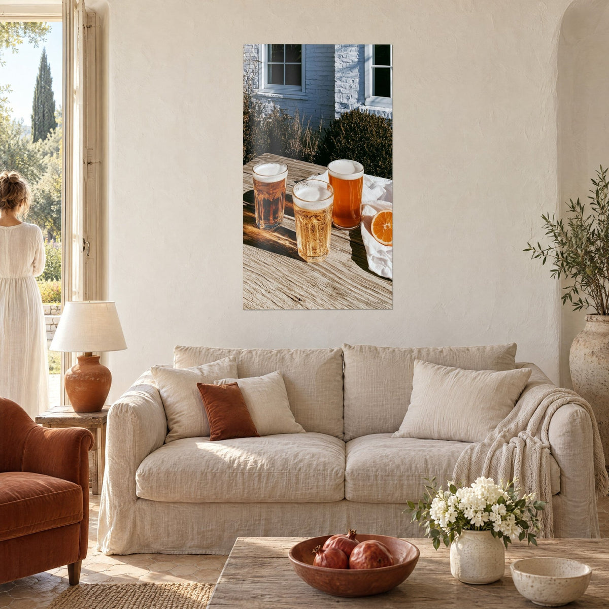 Quadro tre bicchieri di birra spumosa su tavola di legno, arancio e ambra, donna di schiena, sopra un divano in un soggiorno luminoso