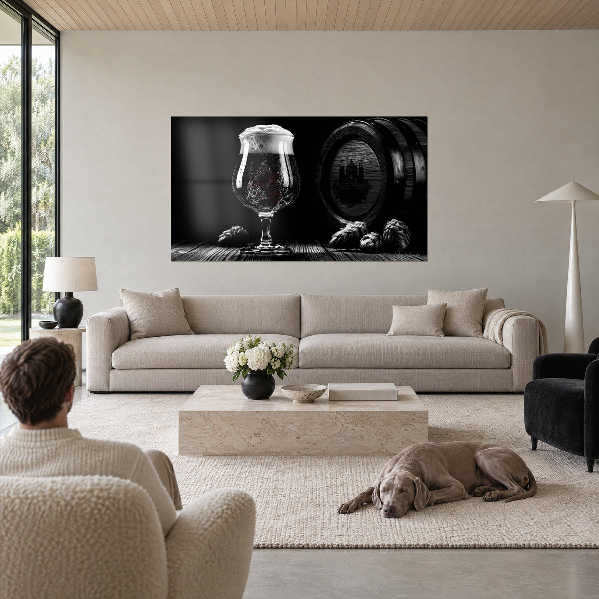 Quadro boccale da birra su piede in bianco e nero, schiuma cremosa traboccante, luppolo e botte di rovere, uomo di spalle e un Weimaraner sopra un divano in un soggiorno luminoso