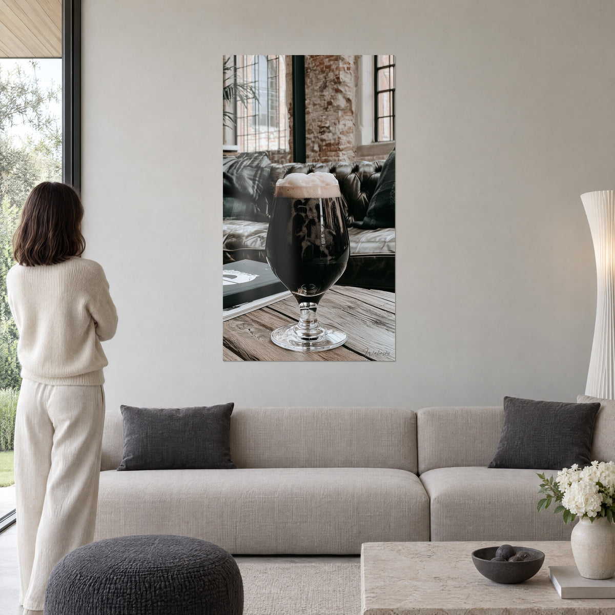 Πίνακας σκούρα μπύρα stout με κρεμώδη αφρό σε ποτήρι με πόδι, καφέ καπιτονέ δερμάτινος καναπές στο βάθος, κρεμασμένος πάνω από καναπέ σε φωτεινό σαλόνι