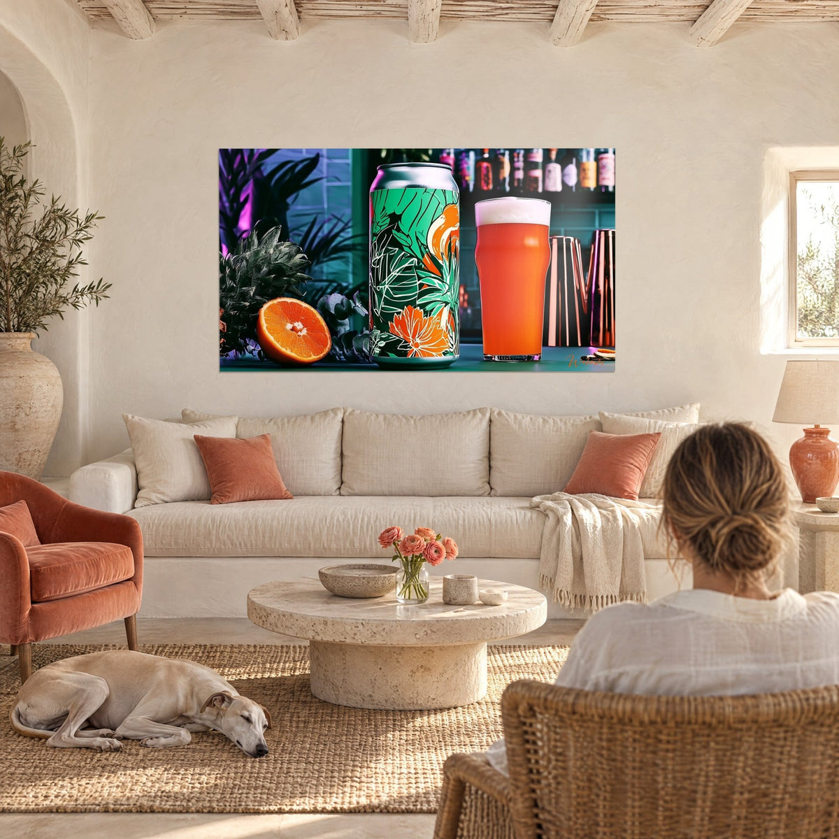 Cuadro con lata de cerveza verde de motivos florales naranjas junto a un vaso de cerveza espumosa, mujer de espaldas y galgo, sobre un sofá en un salón luminoso
