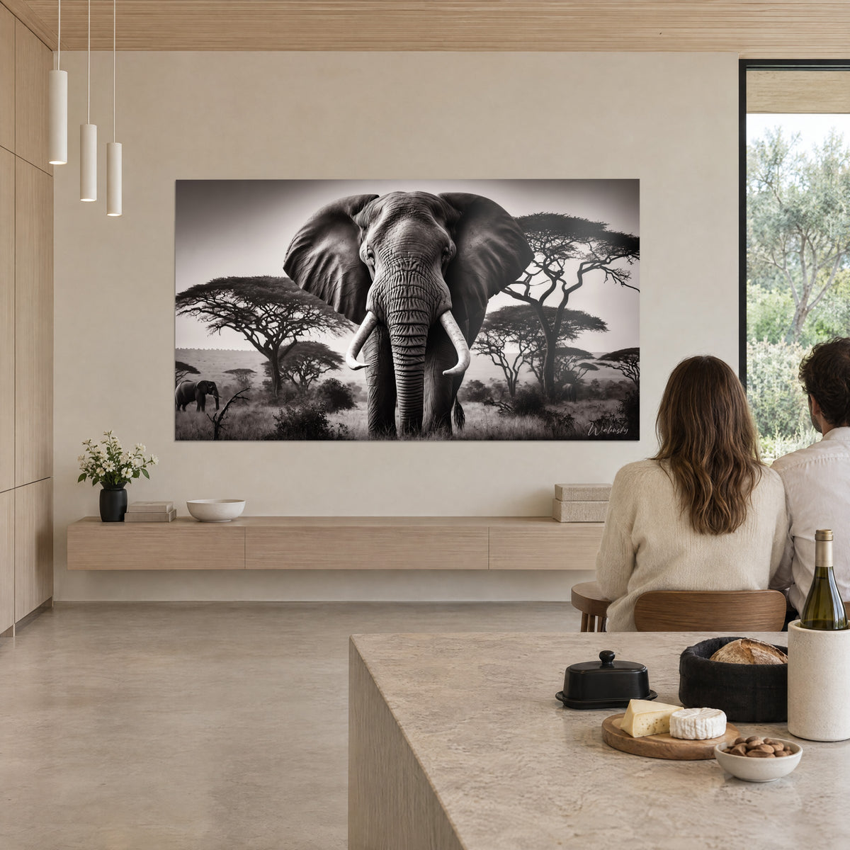 Cuadro de elefante gigante de colmillos blancos, acacias oscuras sobre cielo crema, admirado por una pareja ante una mesa de madera clara