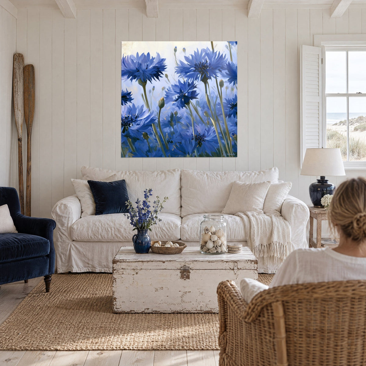 Quadro fiordalisi blu reale con steli verdi, petali frangiati su sfondo crema, donna di spalle sopra un divano in un soggiorno luminoso