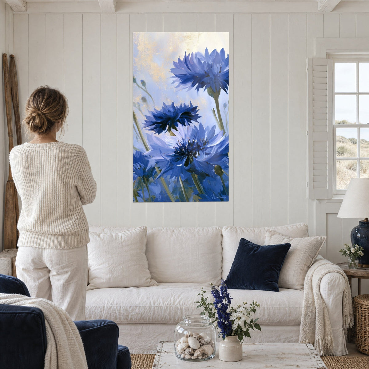 Gemälde Kornblumen mit aufgefächerten indigoblauen Blütenblättern auf goldgetöntem Elfenbeinhintergrund, Frau von hinten, über einem Sofa in einem hellen Wohnzimmer