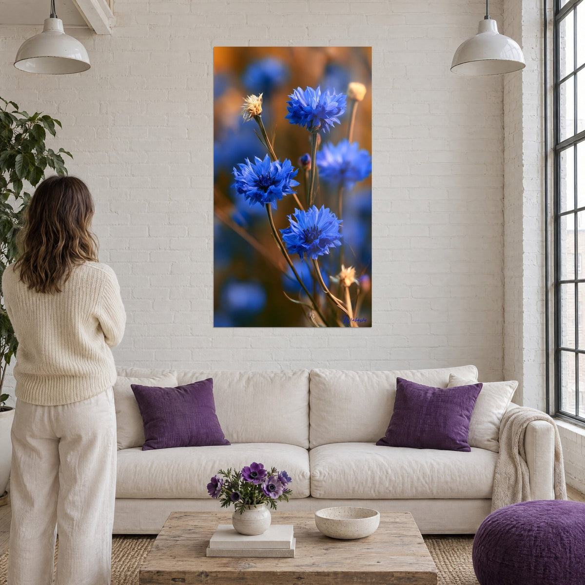 Quadro fiordalisi blu vivo su sfondo ambra, steli piegati, donna di spalle, sopra un divano in un soggiorno luminoso