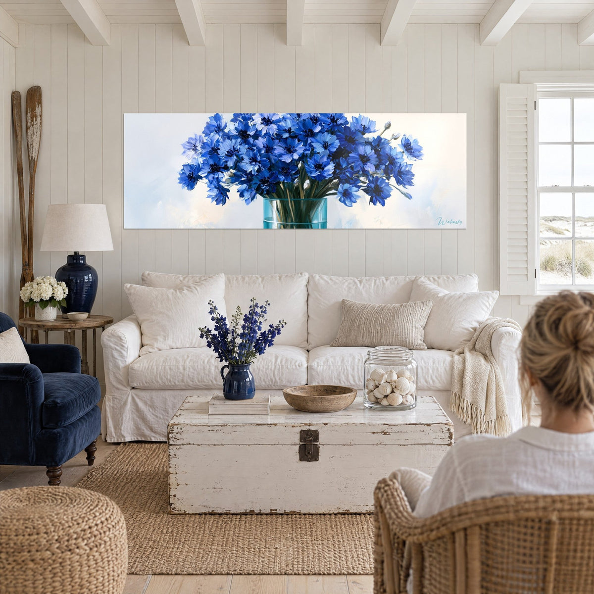 Tableau bouquet de bleuets épanouis, pétales bleu roi et bleu ciel, vase de verre turquoise, au-dessus d'un canapé dans un salon lumineux