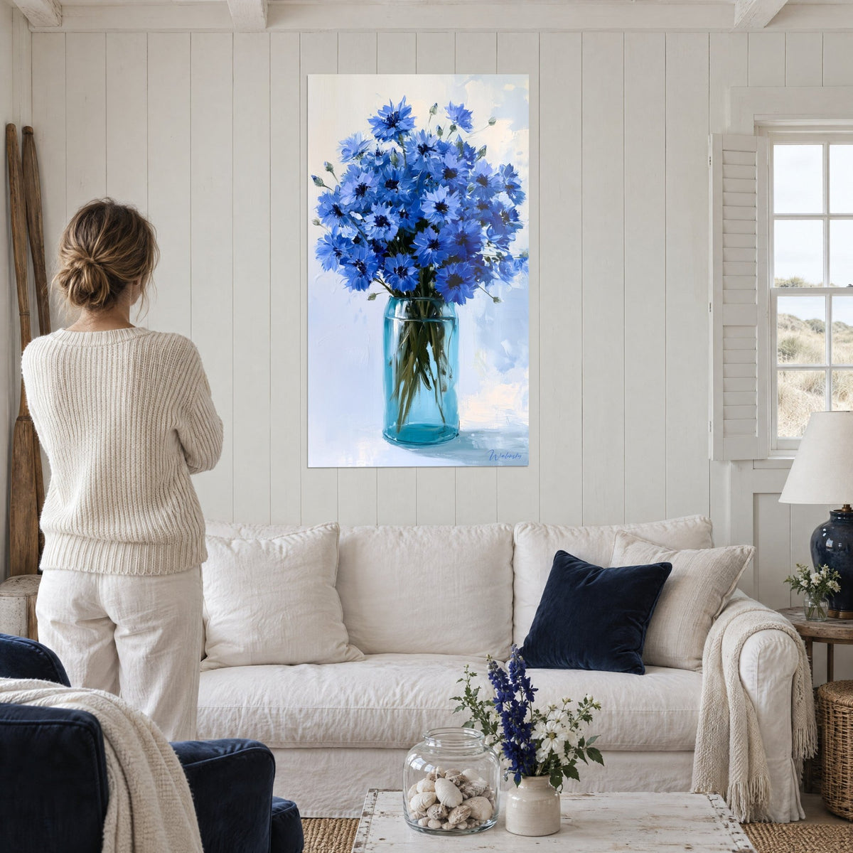 Tableau bouquet de bleuets cobalt dans un bocal turquoise, fond blanc cassé, femme de dos, au-dessus d'un canapé dans un salon lumineux