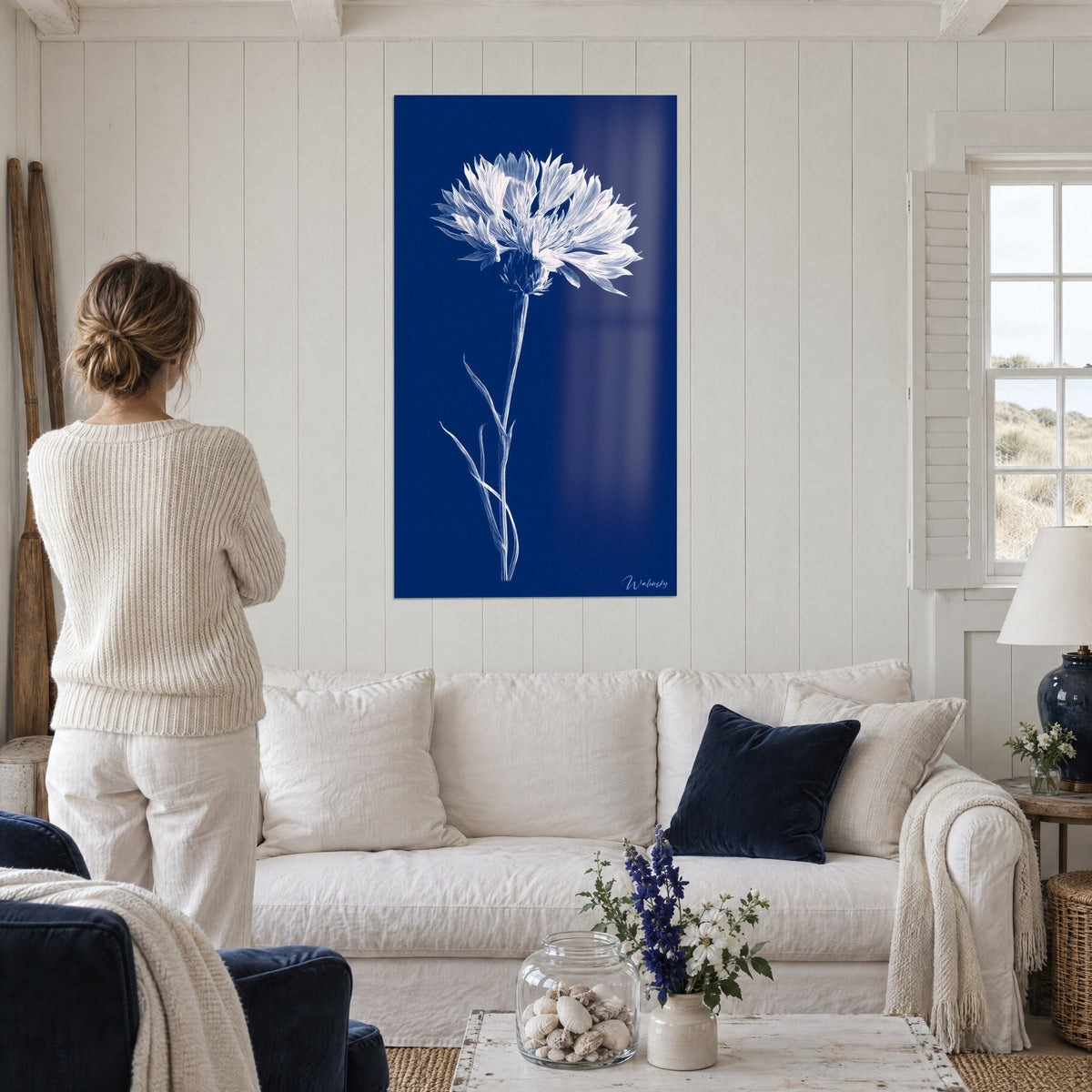 Gemälde Kornblume weiß fein graviert auf marineblauem Grund, Frau im cremefarbenen Pullover von hinten, über einem Sofa in einem hellen Wohnzimmer